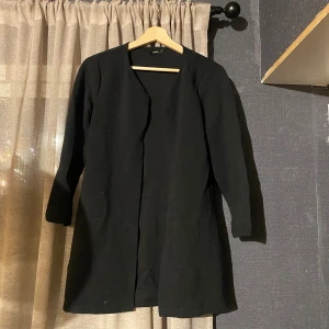 Svart kappa från ONLY i storlek M - Svart lång kappa från ONLY i storlek M. Jackan har en stilren och enkel design utan knappar eller dragkedja, med lång ärm och rak passform. Materialet är mjukt och lätt ribbat vilket ger en snygg struktur. Perfekt för dig som gillar minimalistisk stil.