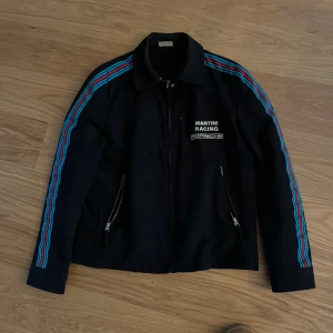 Martini Racing Porsche jacka L - SUPERSKÖNT SVART JACKA. MÅSTE SÄGA ATT DE HAR SMÅ FLÄCKAR SOM EJ GICK BORT I TVÄTTEN. DÄRFÖR DET NEDSATTA PRISET. JACKAN KAN ANDVÄNDAS ISTÄLLET TILL MER SLITSAMMA OUTFITS. Svart jacka från Porsche Design med broderad 'Martini Racing Porsche'-logga på bröstet. Snygga blå och röda ränder längs ärmarna, dragkedja framtill och på fickor. Klassisk krage och sportig vibe, perfekt för dig som gillar racing och streetwear.