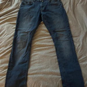 Steve satin Morris jeans - Säljer ett par blå jeans från Morris modell Steve satin med snygga detaljer som broderad logga på fickan och klassiska bakfickor med zigzag-söm. Jeansen har normal passform och är tillverkade i mjukt bomullsmaterial med lite stretch för extra komfort.