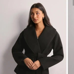 Svart kappa med bälte och bred krage - Snygg svart kappa med bred krage och knytbälte i midjan. Jackan har lång ärm och en stilren, klassisk look som passar perfekt till höst och vinter. Materialet känns mjukt och har en elegant finish. Perfekt för dig som gillar minimalistisk stil.