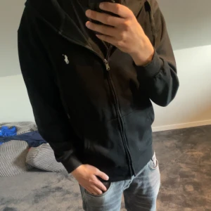 Svart hoodie från Polo Ralph Lauren - Svart hoodie från Polo Ralph Lauren med dragkedja och huva. Klassisk broderad logga på bröstet och stora fickor framtill. Skön passform och perfekt för chill dagar. Snygg och enkel att matcha med jeans eller joggers.