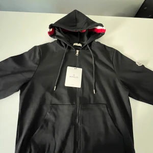 Svart Moncler hoodiejacka med dragkedja - Svart hoodiejacka från Moncler med hel dragkedja, huva med röd och vit rand samt logotyp på ärmen. Jackan har känguruficka framtill och snörning i huvan. Perfekt för dig som gillar stilren och sportig look.