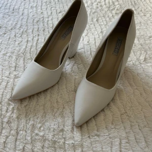 Vita pumps med spetsig tå och klack - Snygga vita pumps från Ex Shoes i storlek 37. Skorna har en spetsig tå och kraftig hög klack som ger en cool siluett. Tillverkade i syntetmaterial med slät finish. Perfekta för dig som gillar stilrena och trendiga skor med lite edge.