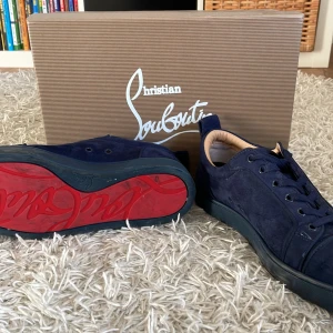 Christian Louboutin blå mocka sneakers - Snygga sneakers från Christian Louboutin i mörkblå mocka med ikonisk röd sula och broderad logga på hälen. Skorna har rund tå, platt sula och klassisk snörning. Perfekta för dig som vill sticka ut med exklusiv design och lyxiga material.