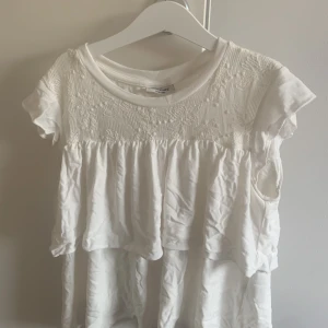 Vit blus med broderad överdel - Supersöt vit blus från kappahl med broderad överdel och volangdetaljer. Blusen har korta ärmar och rund halsringning. Perfekt för en fräsch och romantisk look. Strl 134/140. Lite skrynklig men man kan ju såklart bara stryka den 