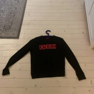 Svart tröja från Hugo Boss med röd logo - Svart långärmad tröja från Hugo Boss med röd HUGO-logga på bröstet. Klassisk rund halsringning och stilren design som passar perfekt till streetwear. Mjuk och skön, perfekt för dig som gillar snygga märkeskläder.