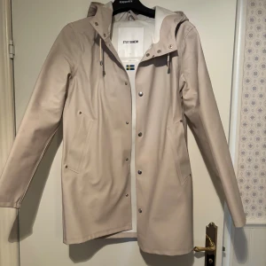 Beige regnjacka/kappa från Stutterheim XS - Snygg beige regnjacka/kappa från Stutterheim i storlek XS. Jackan har huva med snörning, tryckknappar framtill och två fickor. Tillverkad i bomull och polyester med gummerad yta som skyddar mot regn. Perfekt för blöta dagar och stilren look. Mycket bra skick, inga defekter endast använd fåtal gånger!  Nypris 2000kr mitt pris 700kr
