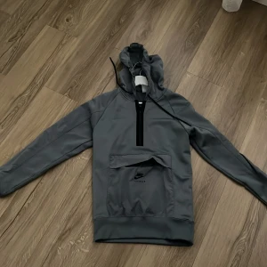 Grå Nike Air Max Therma-Fit hoodie XS - Nike Air Max Therma-Fit hoodie i grått med huva och half zip-dragkedja. Stor känguruficka framtill med Air Max-logga. Tillverkad i mjukt och värmande syntetmaterial, perfekt för kyliga dagar. Långärmad och sportig look.
