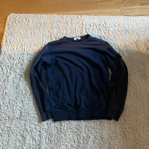 Marinblå sweatshirt från Stone Island - Säljer en stilren marinblå sweatshirt från Stone Island med klassisk rund hals och ikonisk patch på ärmen. Tröjan har långa ärmar och ribbade muddar vid ärmslut och nederkant. Perfekt för dig som gillar streetwear och snygga detaljer. Storlek M/S