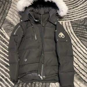 Svart Moose Knuckles dunjacka med päls - Svart dunjacka från Moose Knuckles med vit päls på huvan, flera dragkedjor och snygga detaljer på ärmen. Jackan har hög krage, justerbar huva och logotyp på vänster ärm. Perfekt för kalla dagar med sin tjocka och fluffiga design.