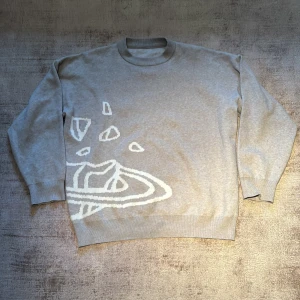 Grå stickad tröja med abstrakt mönster - Säljer en grå stickad tröja med ett coolt vitt abstrakt mönster på framsidan. Tröjan har rund halsringning och långa ärmar, perfekt för dig som gillar en stilren men unik look.