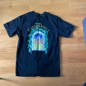 Svart Avenida Casablanca t-shirt - Svart t-shirt från Casablanca med färgglad tryckt grafik på både bröstet och ryggen. Motivet föreställer en byggnad i nattmiljö omgiven av gröna blad och texten 'Avenida Casablanca'. Klassisk passform och rund hals. Perfekt statement-plagg för dig som gillar unika prints. Pris kan diskuteras