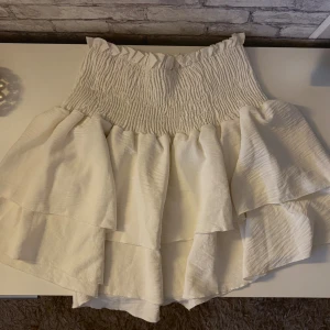 Vit volangkjol  - Supersöt vit kortkjol med bred smock i midjan och flera lager volanger. Kjolen har en luftig känsla och är perfekt för varma dagar. Den är tillverkad i ett lätt tyg som ger ett fint fall och extra volym.