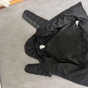 Svart pufferjacka från Adidas - Svart pufferjacka från Adidas med huva och klassiska tre ränder längs ärmarna. Jackan har dragkedja framtill, quiltad design och Adidas-logga på bröstet. Perfekt för kyliga dagar och har en sportig vibe.