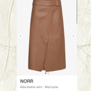 Brun skinnkjol med bälte från NORR - Snygg brun midikjol i äkta lammnappa från NORR. Kjolen har omlott-design, bälte i midjan och en stilren look. Perfekt för dig som vill ha en trendig och tidlös skinnkjol med cool wrap-effekt. På insidan, bredvid dragkedjan finns ett jack i skinnet (se bild). Inga andra anmärkningar. Storlek L, normal storlek. Nypris 2899kr