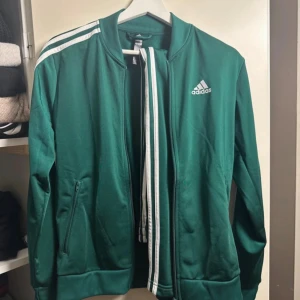 Grön Adidas track jacket med vita ränder - Snygg grön Adidas track jacket med klassiska vita ränder längs ärmarna och dragkedja framtill. Jackan har ribbade muddar, ståkrage och Adidas-logga på bröstet. Perfekt för en sportig och avslappnad stil. Byxor i samma färg finns och detta är ett set. 