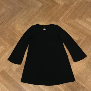 Svart långärmad kortklänning - En stilren svart klänning från Atmosphere med lång ärm och lätt utsvängd passform. Klänningen har en enkel design med liten v-ringning och dekorativ metall-detalj vid halsen. Perfekt för dig som gillar minimalistisk stil.