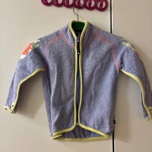 Ljuslila fleecejacka från Molo, stl 110 - Lljuslila fleeceaktig jacka från Molo med gula kantband och coola broderade detaljer på axlarna. Jackan har dragkedja framtill, långa ärmar och tumhål vid ärmsluten. Perfekt för lager-på-lager under höst och vinter. Från djur- rök och parfymfritt hem. 