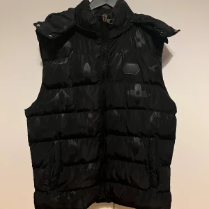 Svart dunväst från Louis Vuitton - Svart dunväst från Louis Vuitton med diskret monogrammönster över hela västen. Västen har hög krage, dragkedja framtill med LV-detalj och två fickor. Tillverkad i 100% polyester och har en puffig, quiltad look. Perfekt för lager-på-lager-stil.