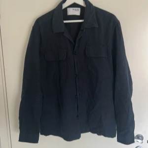 Mörkblå overshirt från Selected Homme - Stilren mörkblå overshirt från Selected Homme med klassisk krage, två bröstfickor med lock och knäppning framtill. Skjortan är långärmad och tillverkad i ett lite grövre tyg som ger en cool och avslappnad look. Använd 2-3 gånger 