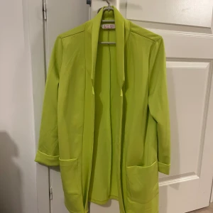Limegrön cardigan kavaj  - Snygg limegrön kavaj från Amisu med öppen front och två stora fickor framtill. Jackan har lång ärm med uppvikta muddar och en rak, avslappnad passform. Perfekt för dig som vill sticka ut med färg och enkel stil.