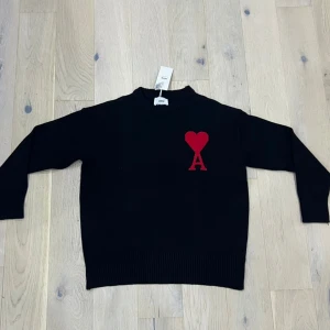 Svart stickad tröja från Ami Paris - Svart stickad tröja från Ami Paris med röd hjärtlogga och bokstaven A på bröstet. Tröjan har rund hals, ribbade muddar och lång ärm. Perfekt för dig som gillar stilrena plagg med en twist.