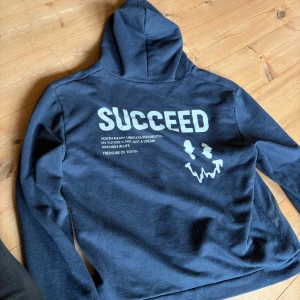 Mörkblå hoodie med tryck 'SUCCEED' - Mörkblå hoodie med stor vit text 'SUCCEED' och coolt ansiktstryck på ryggen. Tröjan har huva och är långärmad, perfekt för chill dagar. Mjuk och skön, troligtvis i bomull eller polyesterblandning. Den fläcken som är på går garanterat bort i tvätten då de är puder 