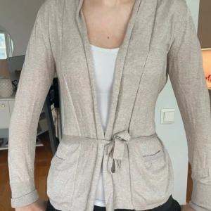 Beige kofta med knytband - Beige kofta med långa ärmar, två fickor framtill och knytband i midjan. Jackan har en mjuk och tunn känsla, perfekt för lager-på-lager. 