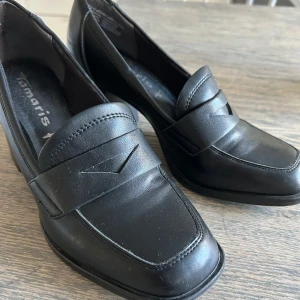 Svarta loafers med klack från Tamaris - Stilrena svarta loafers från Tamaris i skinn med chunky klack och klassisk pennyloafers-detalj framtill. Snyggt minimalistisk design med rund tå och markerad sula, perfekta för dig som vill ha en dressad look med lite edge.