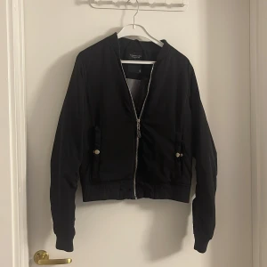 Svart bomberjacka från The Black Label - Svart bomberjacka från The Black Label Collection i storlek S. Jackan har klassisk bombermodell med ribbade muddar, dragkedja framtill och två fickor med knappdetaljer. Snygg och enkel design som passar till många olika stilar.