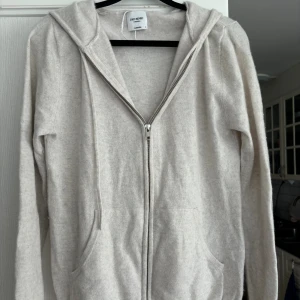 Beige kashmirhoodie från John Henric - Mjuk och lyxig beige zip hoodie i  kashmir från John Henric. Tröjan har dragkedja framtill, huva och två fickor. Perfekt för dig som vill ha en stilren och bekväm look med premiumkänsla.
