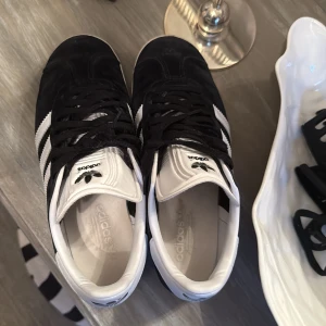 Adidas Gazelle svartvita sneakers - Svartvita Adidas Gazelle sneakers i mocka med klassiska vita ränder och vit hälkappa med logga. Skorna har snörning och platt sula, samt diskret guldfärgad Gazelle-text på sidan. Perfekta för en sportig och stilren look.