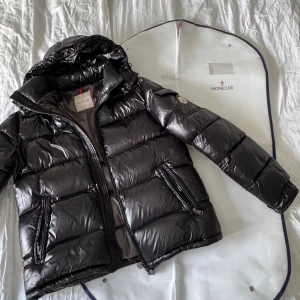  Moncler Maya - Säljer en svart pufferjacka från Moncler med glansig finish och huva. Jackan har dragkedja framtill, två fickor med dragkedja och en ärmficka med Moncler-logga. Insidan har ett unikt serietidningsmärke. Perfekt för kalla dagar!