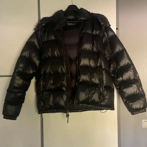 Svart pufferjacka från Polo Ralph Lauren - Säljer en svart, glansig pufferjacka från Polo Ralph Lauren. Jackan har huva, dragkedja framtill och två fickor med dragkedja. Den är vadderad och har en oversized look, perfekt för kalla dagar. Materialet är polyester och jackan är riktigt mjuk och skön.