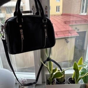 Svart handväska från Misako - Snygg svart handväska från Misako med två silverfärgade dragkedjor framtill och avtagbar axelrem. Väskan är tillverkad i syntetmaterial med en stilren, boxig form och har praktiska handtag. Perfekt storlek för att få plats med allt viktigt. Dragkedjan har tyvärr gått sönder men den går kanske att laga men det är inget jag har hunnit göra. Hör gärna av er kring priser osv💗