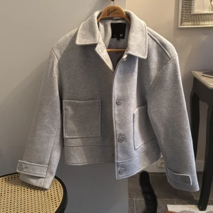 Ljusgrå ulljacka från 157, stl M - Snygg ljusgrå jacka från 157 i ullmix med klassisk krage, stora framfickor och knäppning med knappar. Jackan har en boxig, loose passform och långa ärmar med knappdetalj vid ärmslut. Perfekt för dig som gillar stilren och enkel design.