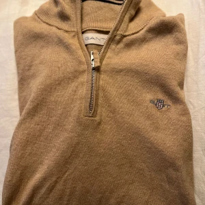 Beige stickad tröja från Gant stl 152 - Snygg beige stickad tröja från Gant i storlek 152 med halv dragkedja och broderad logga på bröstet. Tröjan har lång ärm och klassisk passform, perfekt för lager-på-lager. Mjuk och skön känsla