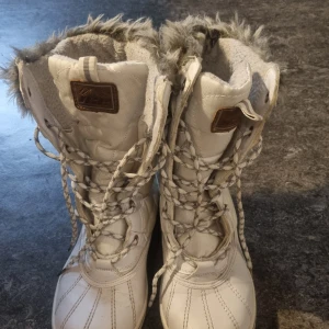 Vita vinterboots från ICE med fuskpäls - Säljer ett par vita vinterboots från ICE med snörning och mysig fuskpäls upptill. Skorna är vattenavvisande och har en robust platt sula. Materialmix av syntet och skinn, perfekt för kalla dagar. Snygga detaljer med logopatch och mönstrade skosnören. Observera slitage på dom sista 3 bilderna.