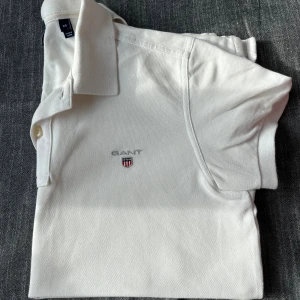 Vit slim fit piké från Gant XS - Vit pikétröja från Gant i slim fit-modell med klassisk krage och korta ärmar. Broderad Gant-logga med sköld på bröstet. Tillverkad i mjuk bomull som känns skön mot huden. Perfekt för en clean och stilren look.