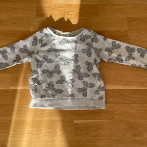Grå tröja med fjärilsmönster - Superfin ljusgrå tröja med svarta fjärilar över hela tyget. Tröjan har rund halsringning och långa ärmar. Mjuk och skön i bomullsmaterial, perfekt för dig som gillar söta detaljer och vill ha något bekvämt och stilrent.