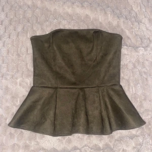 Olivgrön peplum tubtopp - Säljer en olivgrön tubtopp med peplumvolang nertill. Toppen är axelbandslös och har en snygg, figurnära passform. Materialet känns som mocka och ger en lyxig känsla. Perfekt för dig som vill sticka ut med en unik och trendig topp. Endast använd en gång, därför säljer jag den!