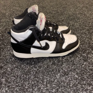 Nike Dunk High svartvita sneakers - Säljer ett par Nike Dunk High sneakers i svart och vitt skinn med klassisk svart swoosh och svart snörning. Skorna har hög siluett, perforerad tå och vit sula. Tungan har Nike-logga i rött. Perfekta för dig som gillar streetstyle och vill sticka ut.     I VERKLIGHETEN SER SKORNA UT I BÄTTRE SKICK 