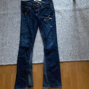 Mörkblå bootcut jeans från Hollister - Säljer ett par mörkblå jeans från Hollister med bootcut passform. Jeansen har slitna detaljer framtill och klassiska bakfickor med broderade sömmar. Tillverkade i jeansmaterial med låg midja och snygg tvätt.