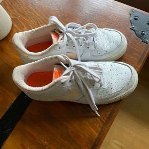 Nike Air Force 1 vita sneakers - Fräscha Nike Air Force 1 sneakers i vitt skinn med ljusbeige swoosh och orange detaljer på insidan och hälen. Klassisk rund tå, snörning och platt sula. Perfekt för dig som gillar stilrena och tidlösa sneakers med en twist. Stl 37,5 💓💓💓