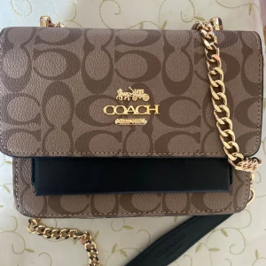 Coach väska - Snygg axelväska från Coach i beige och svart med klassiskt logomönster. Väskan har guldfärgad kedja, detaljer i svart skinn och Coach-emblem i guld på framsidan. Perfekt storlek för det viktigaste och riktigt trendig design.