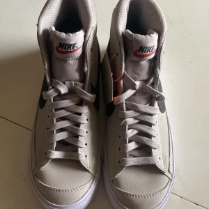 Nike Blazer Mid sneakers beige - Nike Blazer Mid sneakers i beige med svart Swoosh och vit sula. Klassisk hög modell med snörning och retrovibbar. Ovandel i syntet och skinn, detaljerad med Nike-logga på plös och häl. Perfekta för dig som gillar stilrena och tidlösa sneakers.