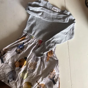 Grå långärmad klänning med fågelmönster från Molo - Supersöt klänning från Molo med ribbad grå överdel och långärmade ärmar. Kjolen är ljusgrå med ett detaljerat fågelmönster i olika färger som ger en cool och lekfull vibe. Tillverkad i mjuk bomull, perfekt för dig som gillar unika prints och bekväma plagg.