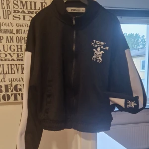 Svart och vit bomberjacka FB Sister XS - Svart och vit bomberjacka från FB Sister i storlek XS. Jackan har dragkedja framtill, broderad text och motiv på bröstet samt ärmen. Sportig look med vita paneler längs ärmarna och elastisk nederkant. Perfekt för en streetstyle vibe.