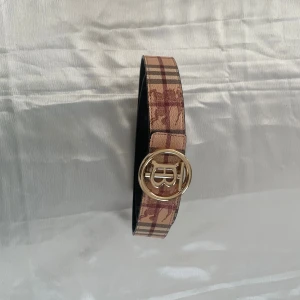 Burberry rutig skärp med guldspänne - Snyggt Burberry-bälte i klassiskt beige och brunt rutigt mönster med ikoniskt TB-guldspänne. Bältet är gjort i skinn och har svarta kanter. Perfekt accessoar för att lyfta din outfit med en lyxig touch.