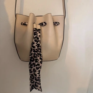 Beige axelväska med leoparddetalj - Snygg beige axelväska i syntetmaterial med justerbart axelband och coolt leopardmönstrat band framtill. Väskan har dragskostängning och en stilren bucket-form som ger en trendig vibe.